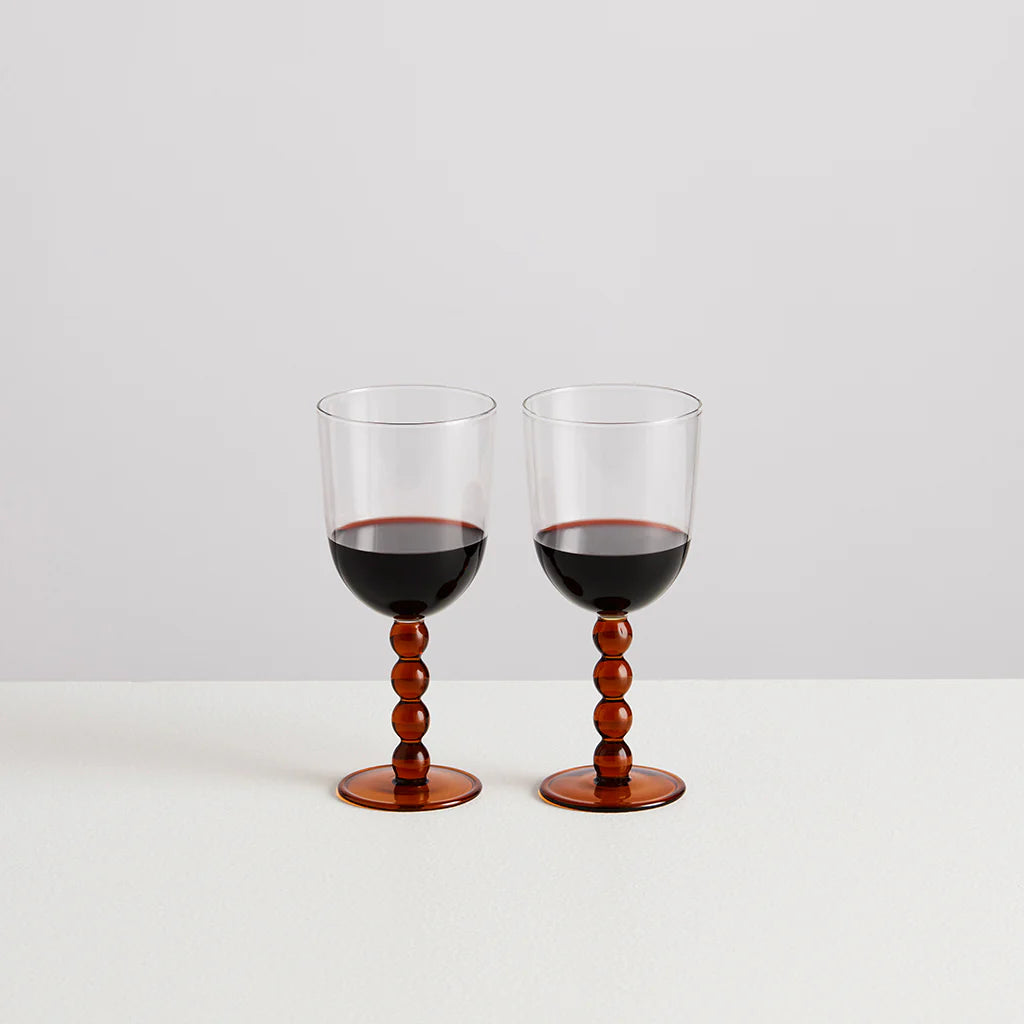 Two wine glasses filled with red wine. The body of the glasses are clear and the stems and base are formed in amber baubles