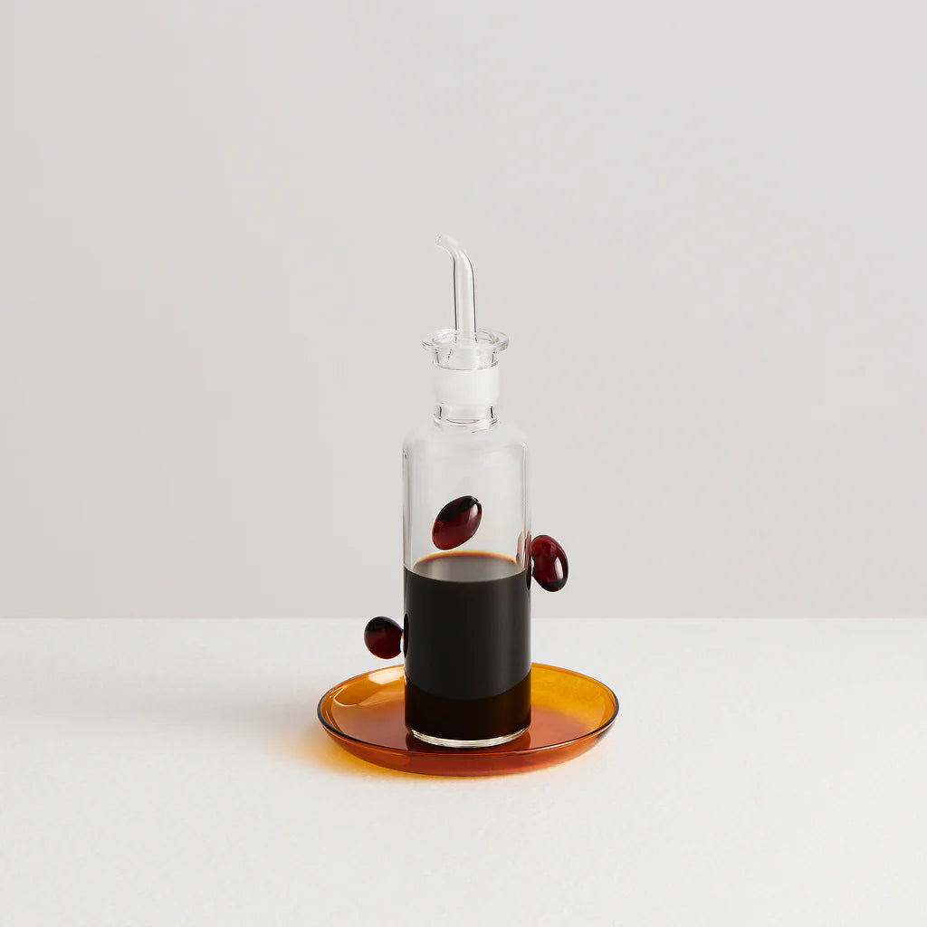 A glass bottle decorated with glass olives and filled with balsamic vinegar. The bottle is placed on an amber coloured glass plate