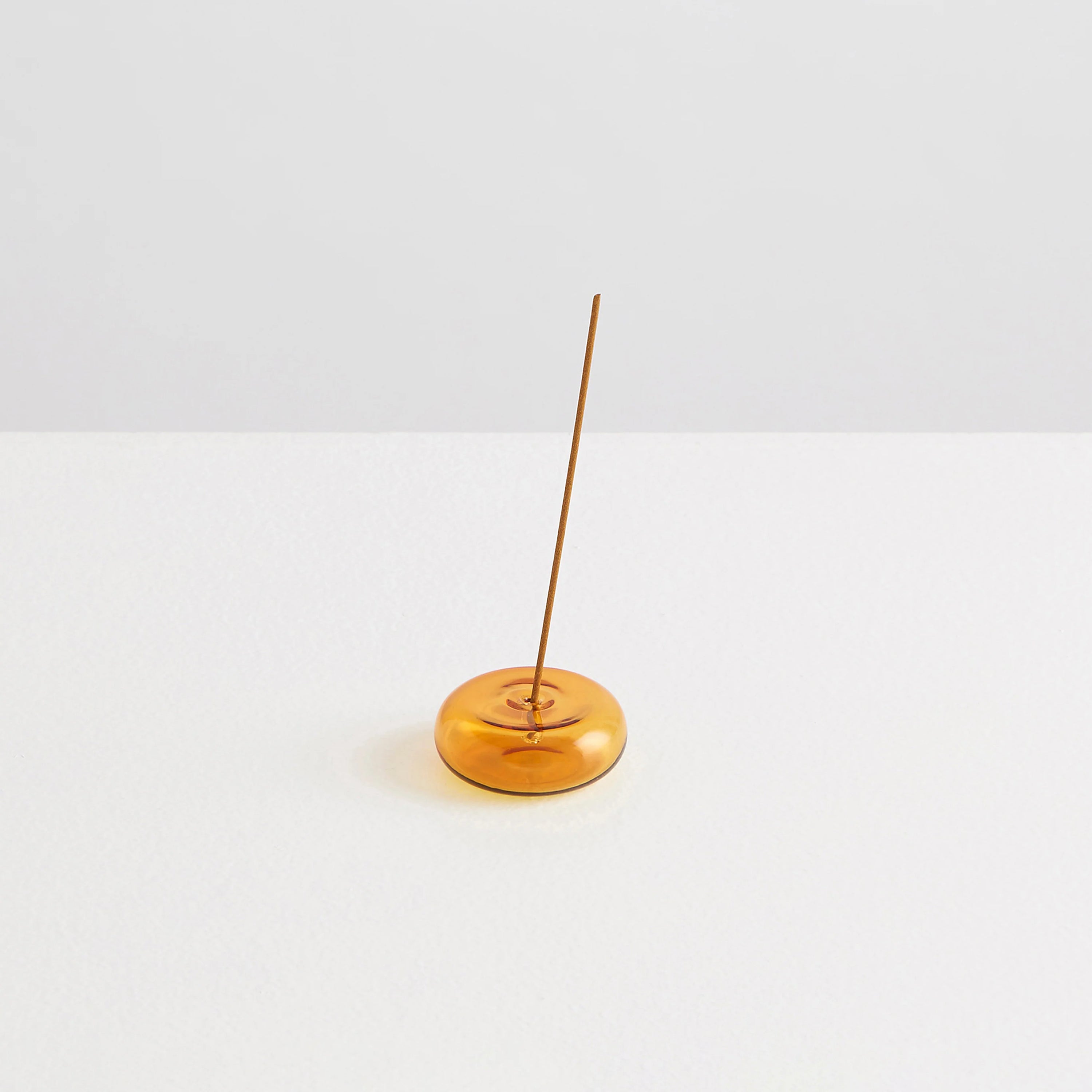 A small pebble shaped clear amber glass incense holder with a stick of incense on a white background