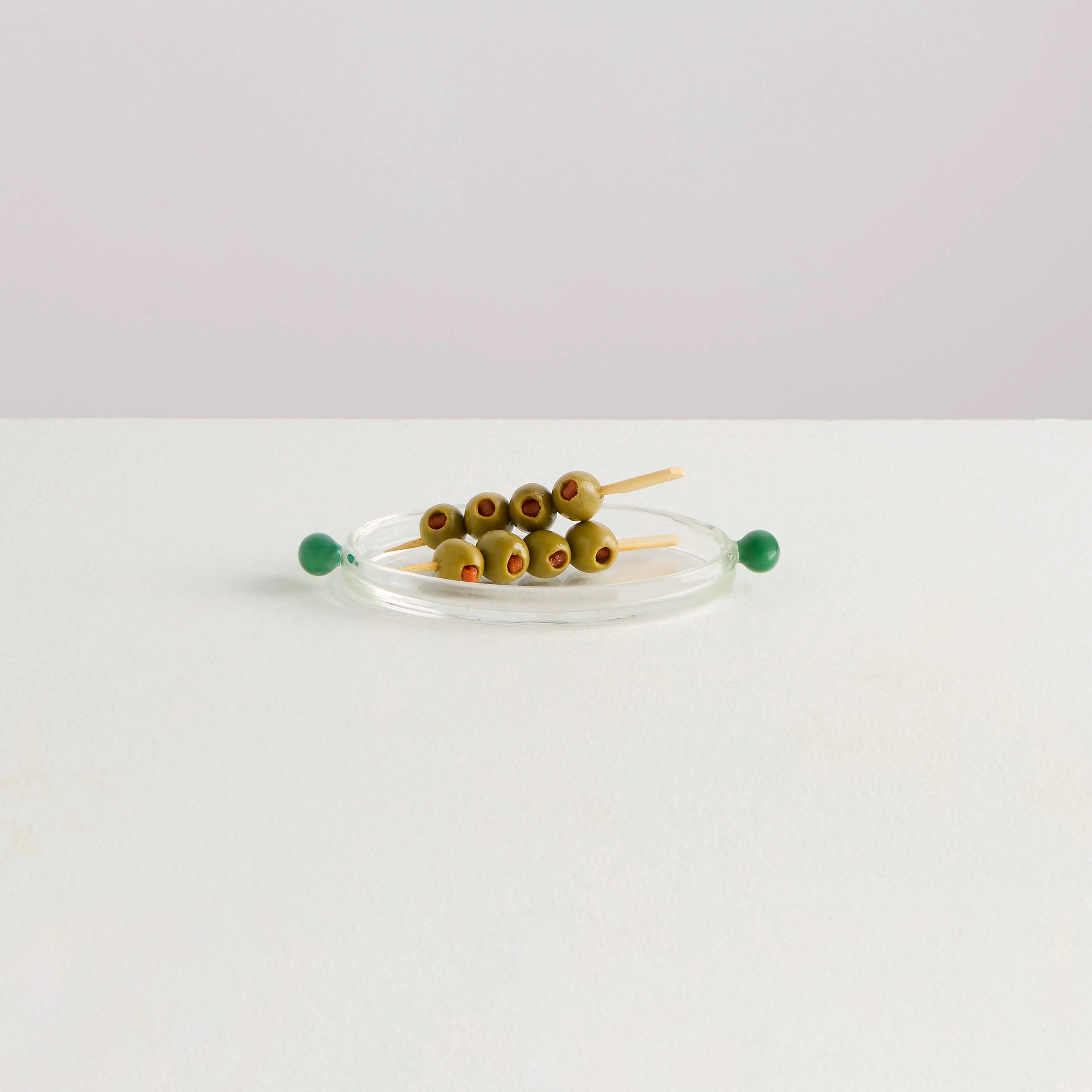 A clear glass platter with two olive shaped green handles on either side with a platter of skewered green olives