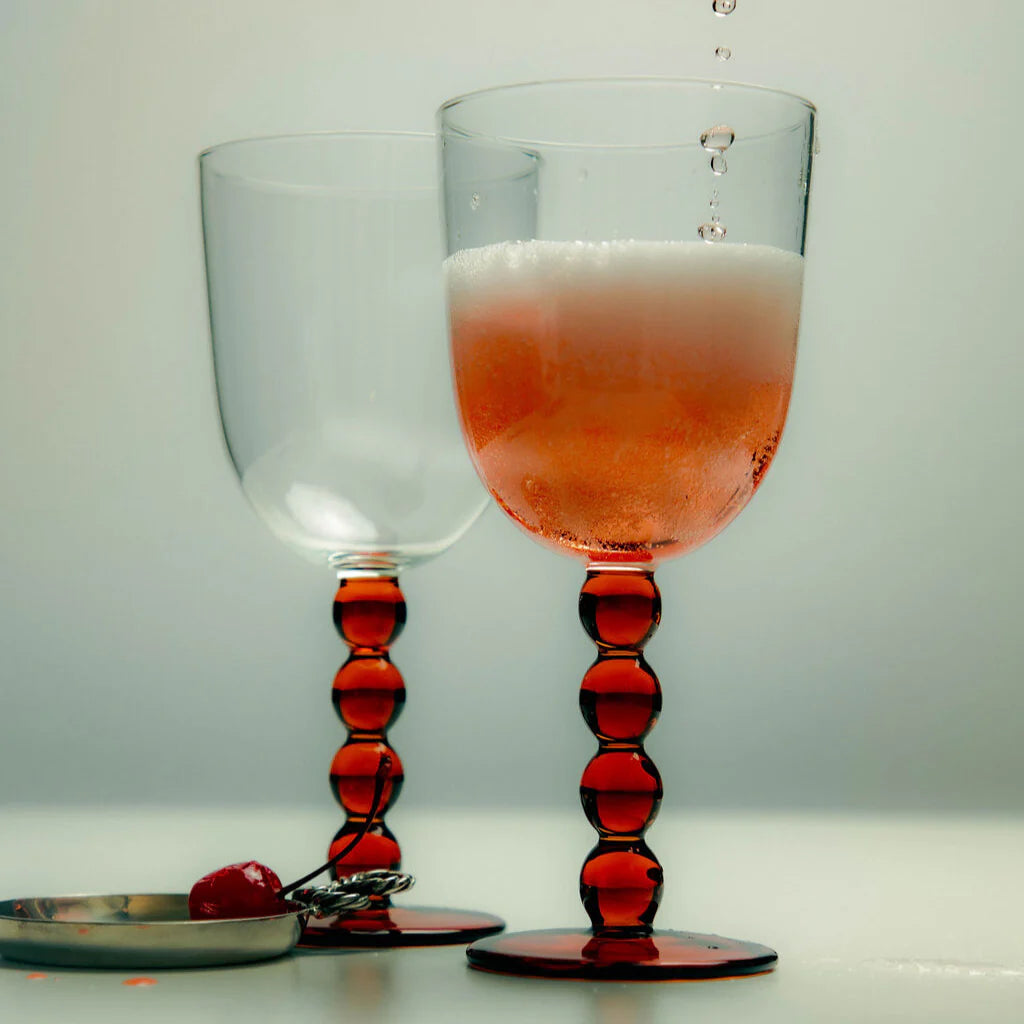 Two wine glasses one empty and the other filled with a sparkling rosé wine. The body of the glasses are clear and the stems and base are formed in amber baubles. These are set in a stylised able setting alongside a silver dish holding a maraschino cherry.