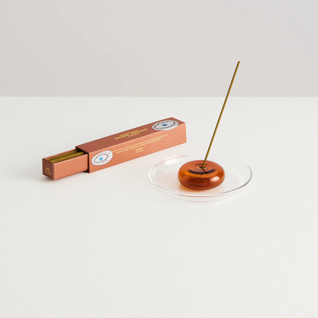 A gift set on a white background. Depicting an opened amber coloured box of incense. Beside the incense box is a pebble shaped clear glass incense holder holding a stick of incense and sat upon a small clear glass plate.