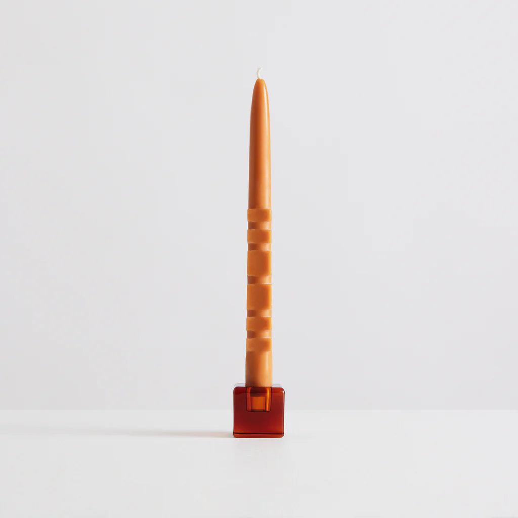 A glass amber cube shaped candle holder holding a geometrically textured beeswax candle. 