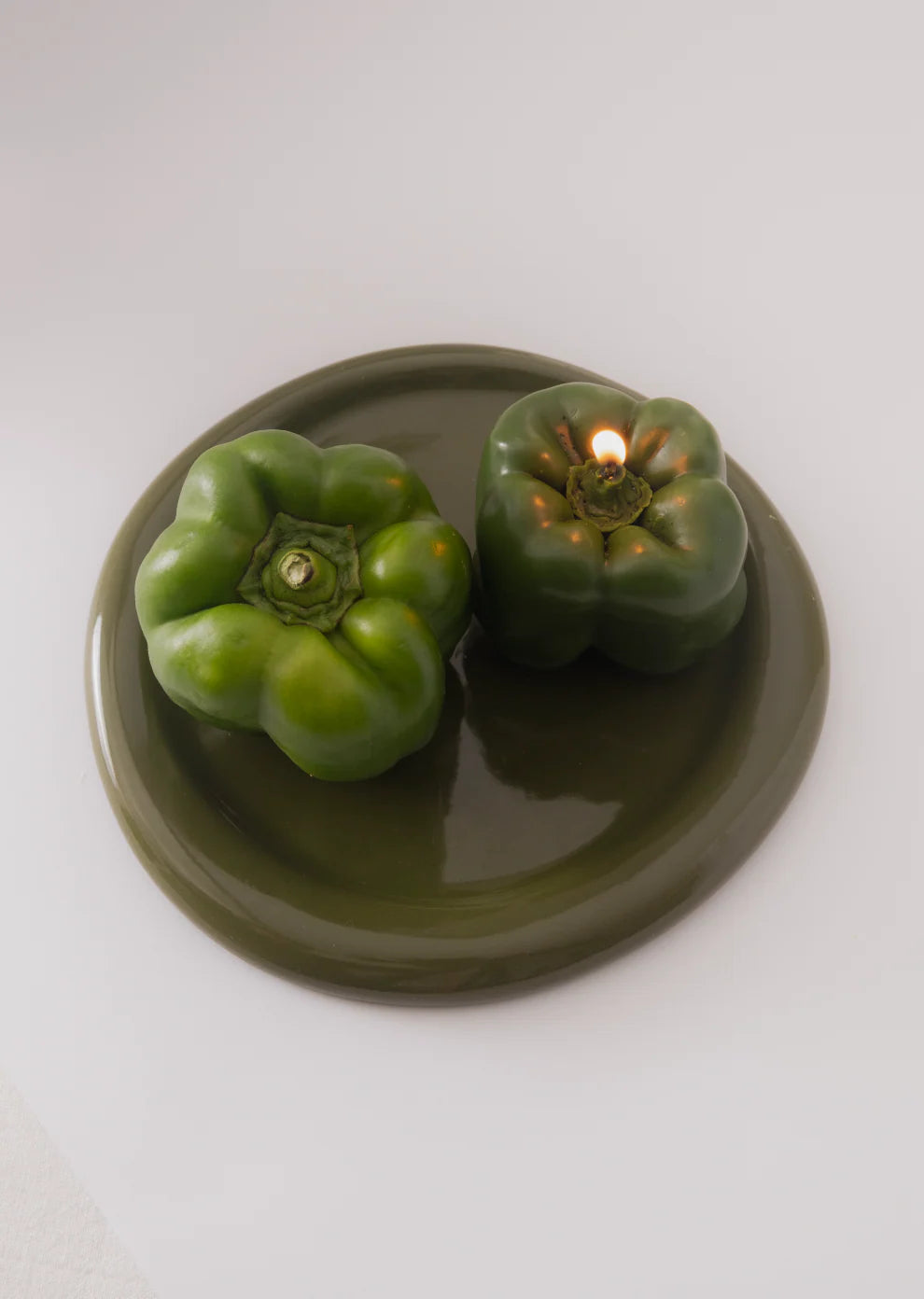 Two green capsicum shaped candles on a round green plate. The candle on the right hand side is lit