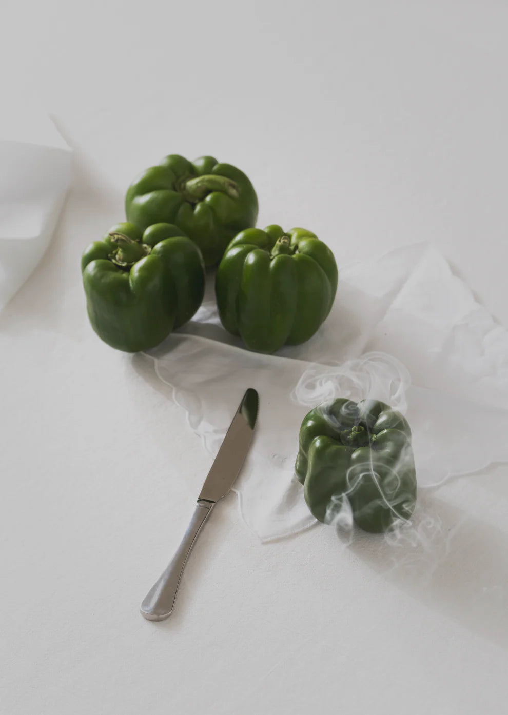 Four green capsicum shaped candles on a table spread with two cotton serviettes and a dinner knife. The flame in the candle closest to the bottom has just been extinguished so smoke rises from this bottom candle