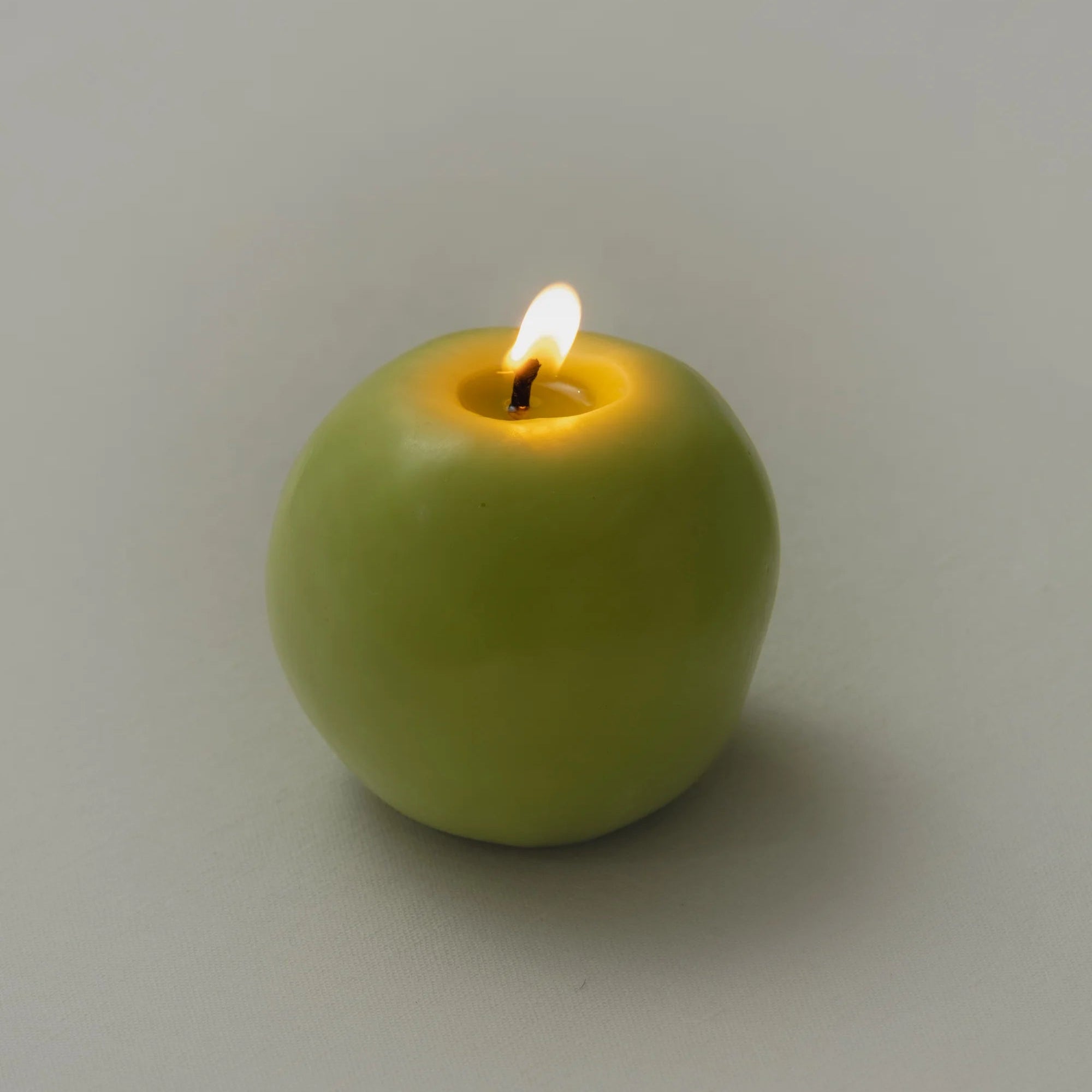 A green apple shaped candle with a lit flame on a moody coloured background